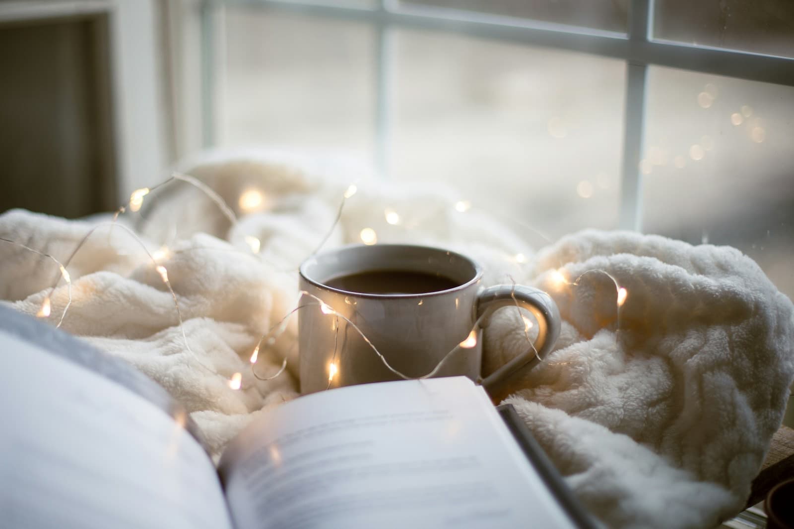 An open book and a mug of coffee on a soft blanket by a window, with small string lights.