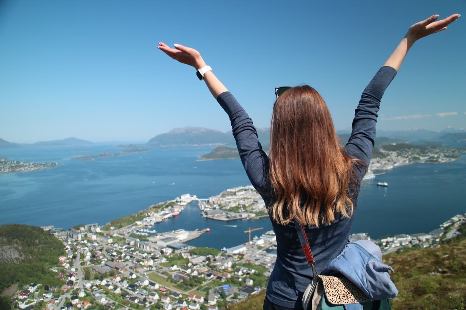 A young woman, seen from behind, throws both arms up on a hilltop above a sunny harbor town and blue fjord.