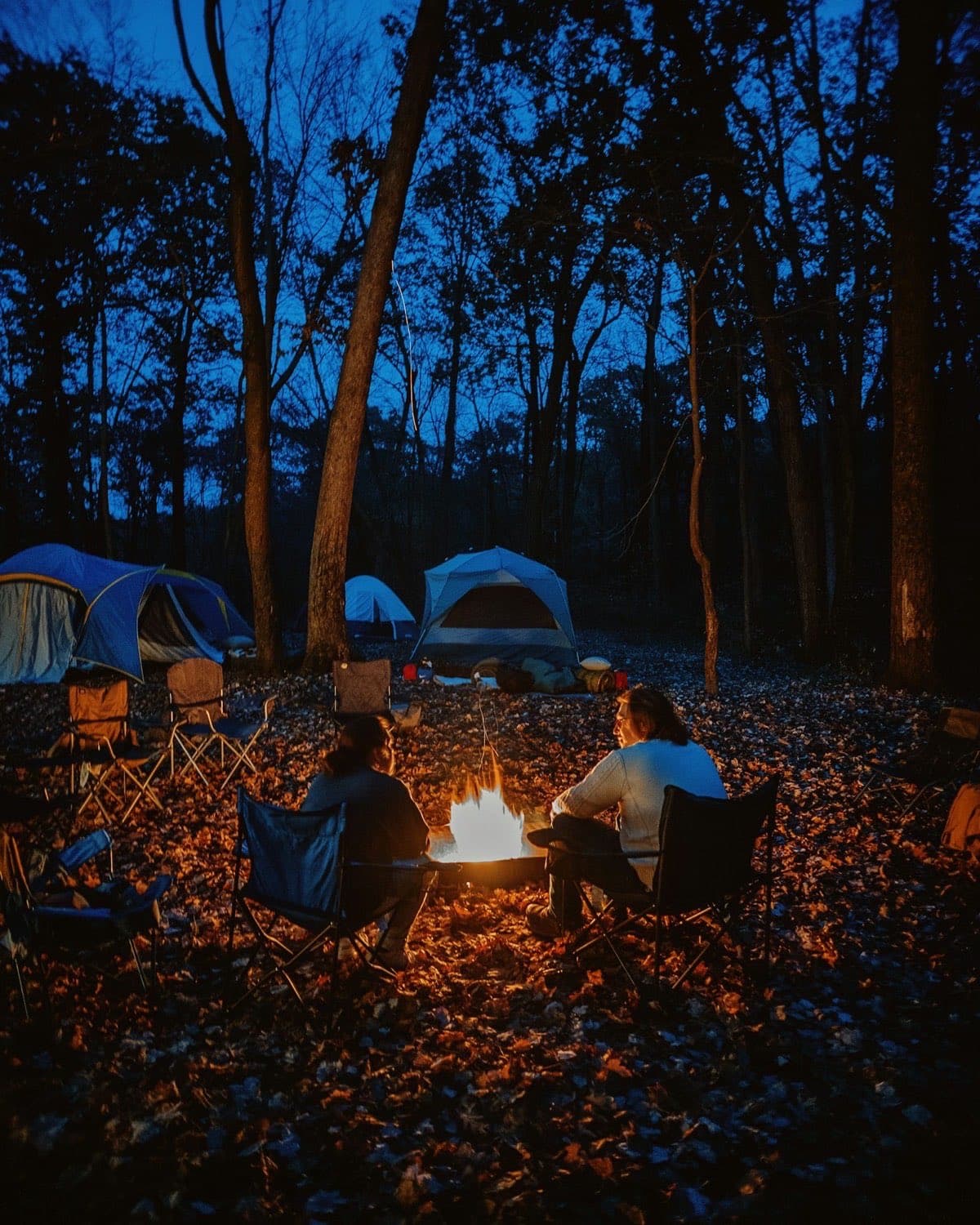 Two people, seen from behind, talking in camp chairs beside a small campfire in an autumn forest at dusk, with tents among the trees.