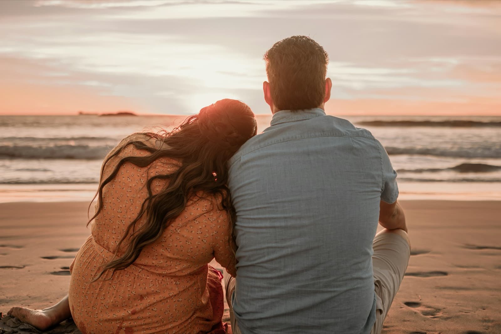 A young couple, seen from behind, sitting close together on the sand and watching the sun set over the waves.