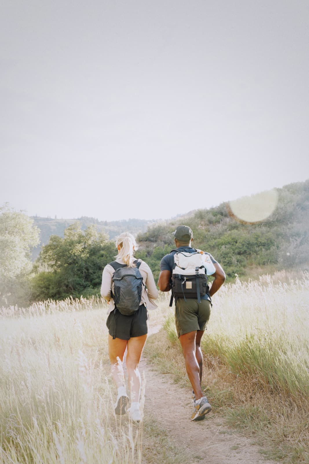 A young couple with small backpacks, seen from behind, jogging side by side along a dirt trail through tall golden grass.