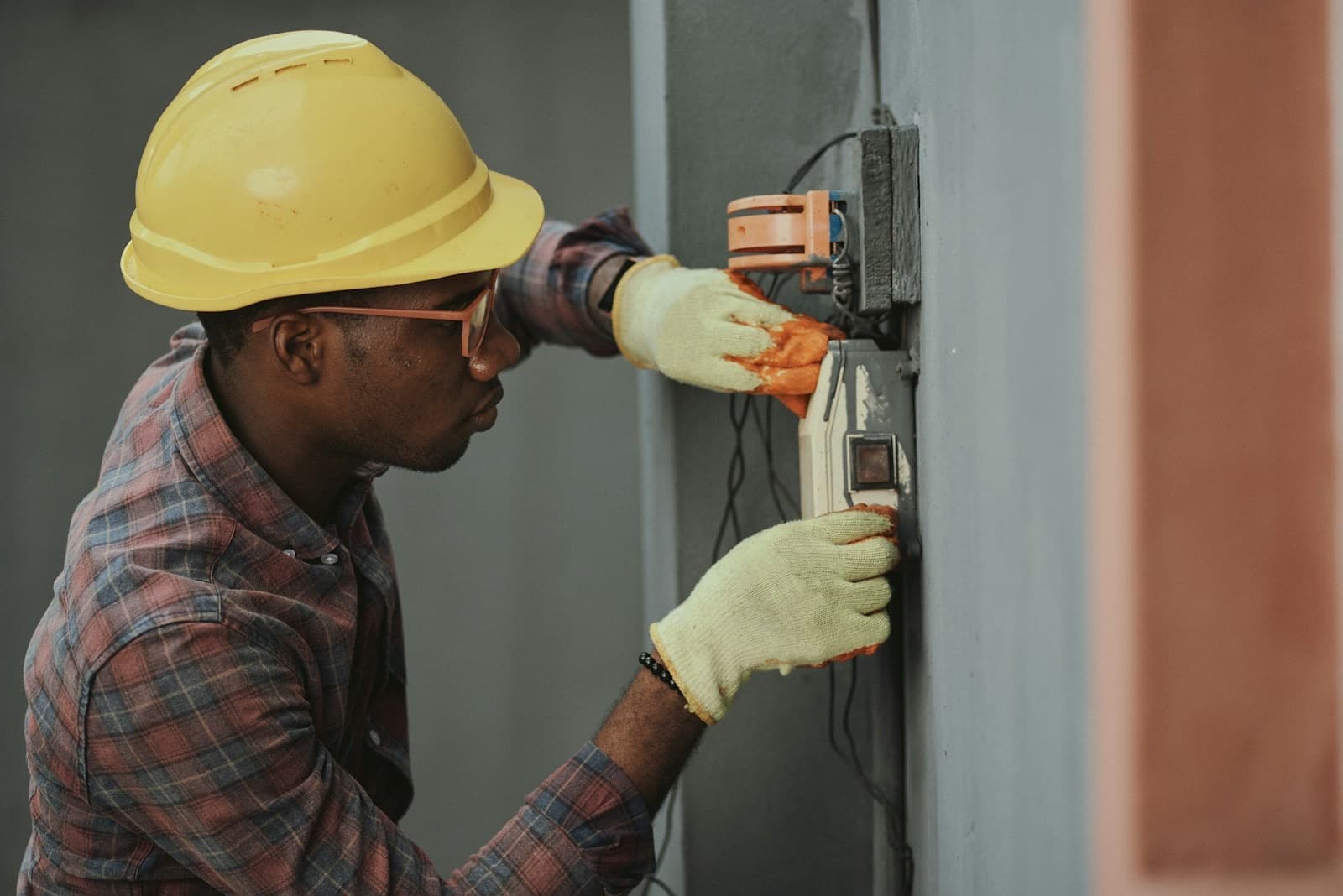 An electrician in a yellow hard hat and work gloves wiring an electrical box.