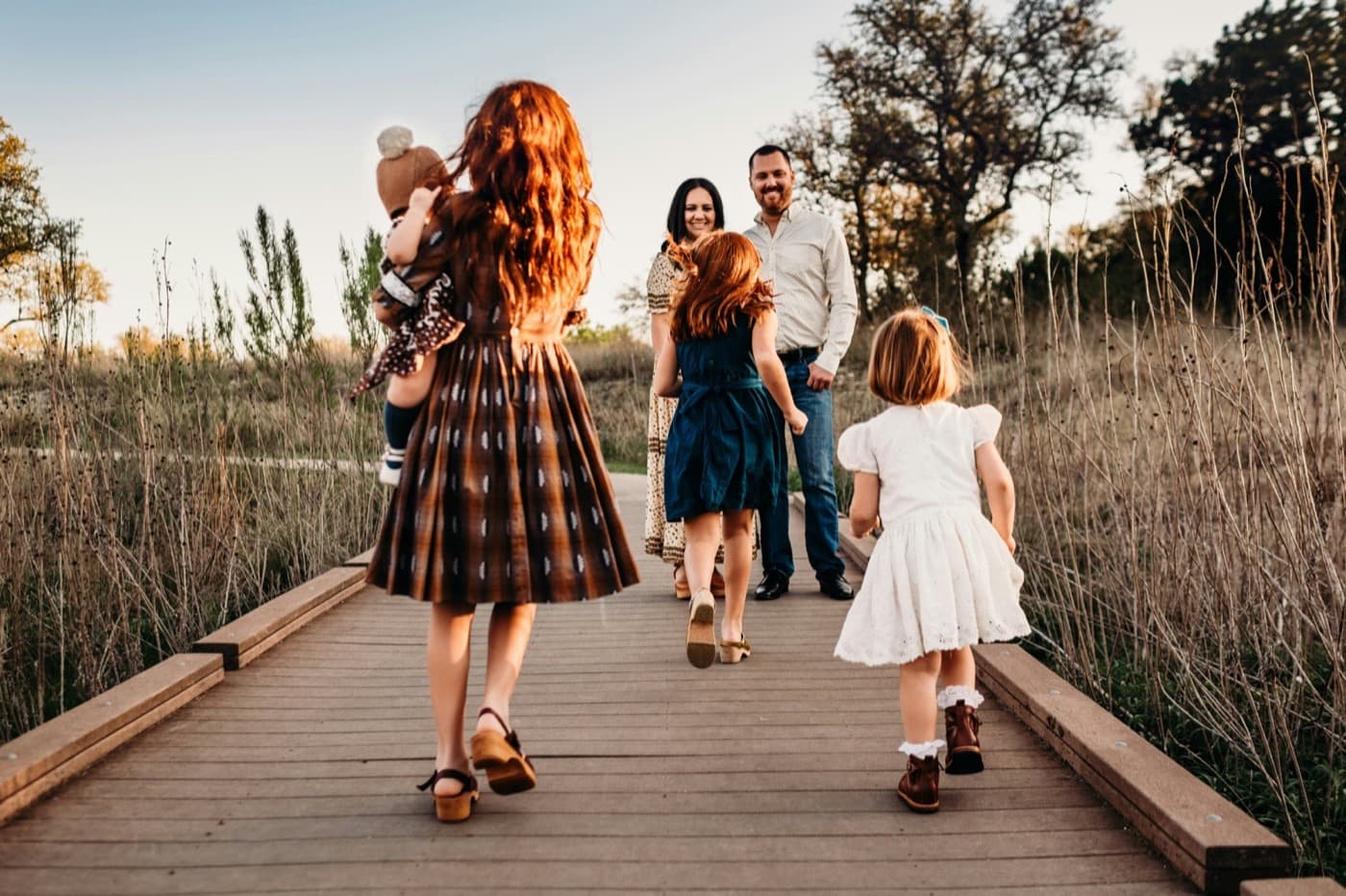 Three young girls, the eldest carrying a baby, running along a wooden boardwalk toward their smiling parents at golden hour.