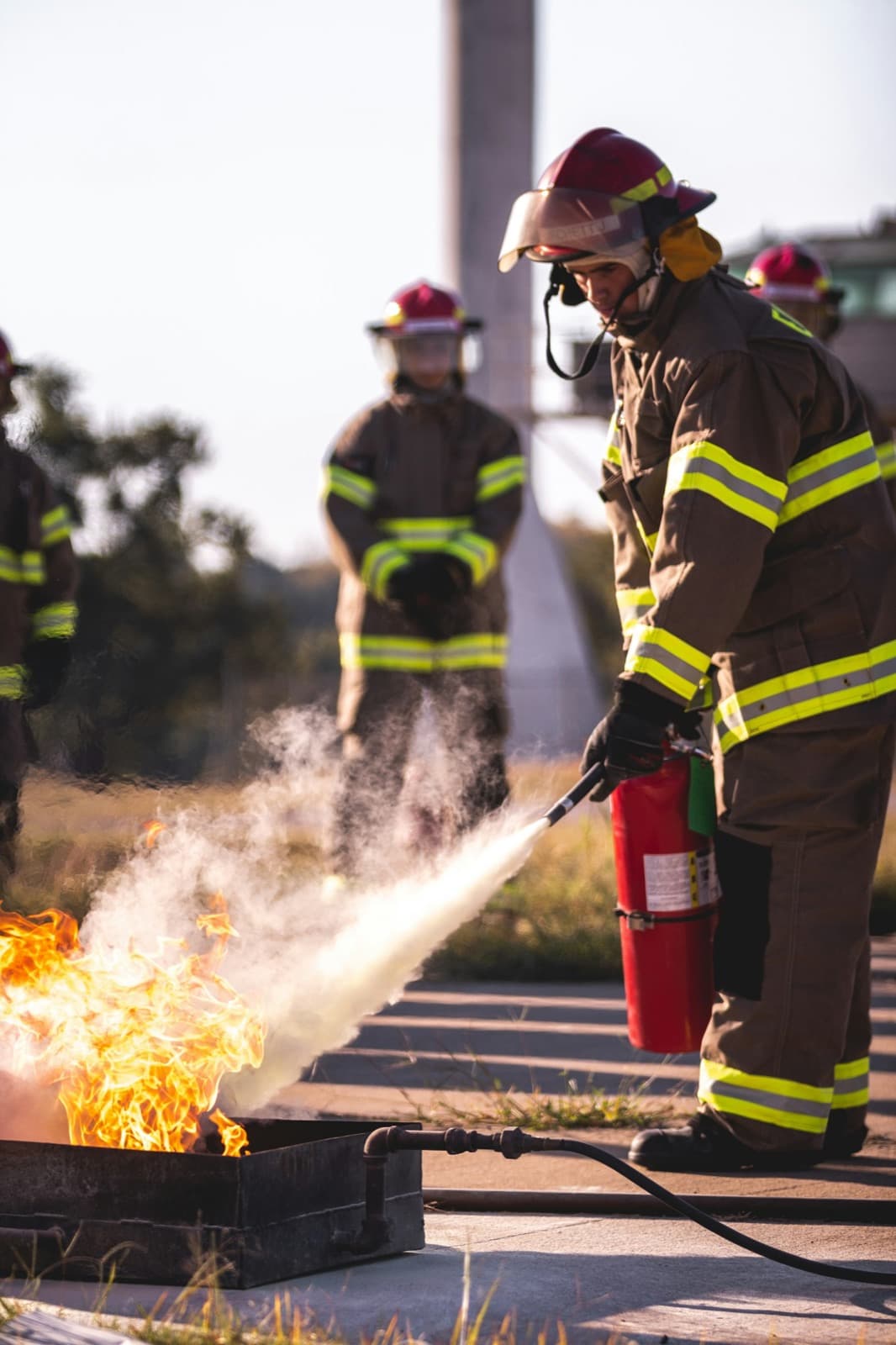 A firefighter in full gear putting out a training fire with an extinguisher while others look on.