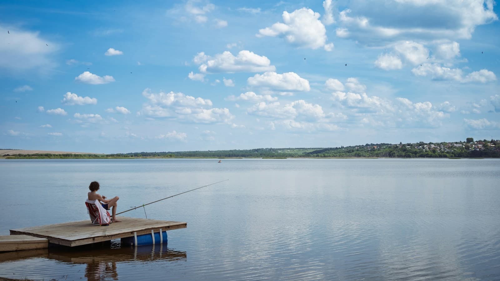 A person fishing from a small dock on a wide, calm lake under a sky full of clouds.