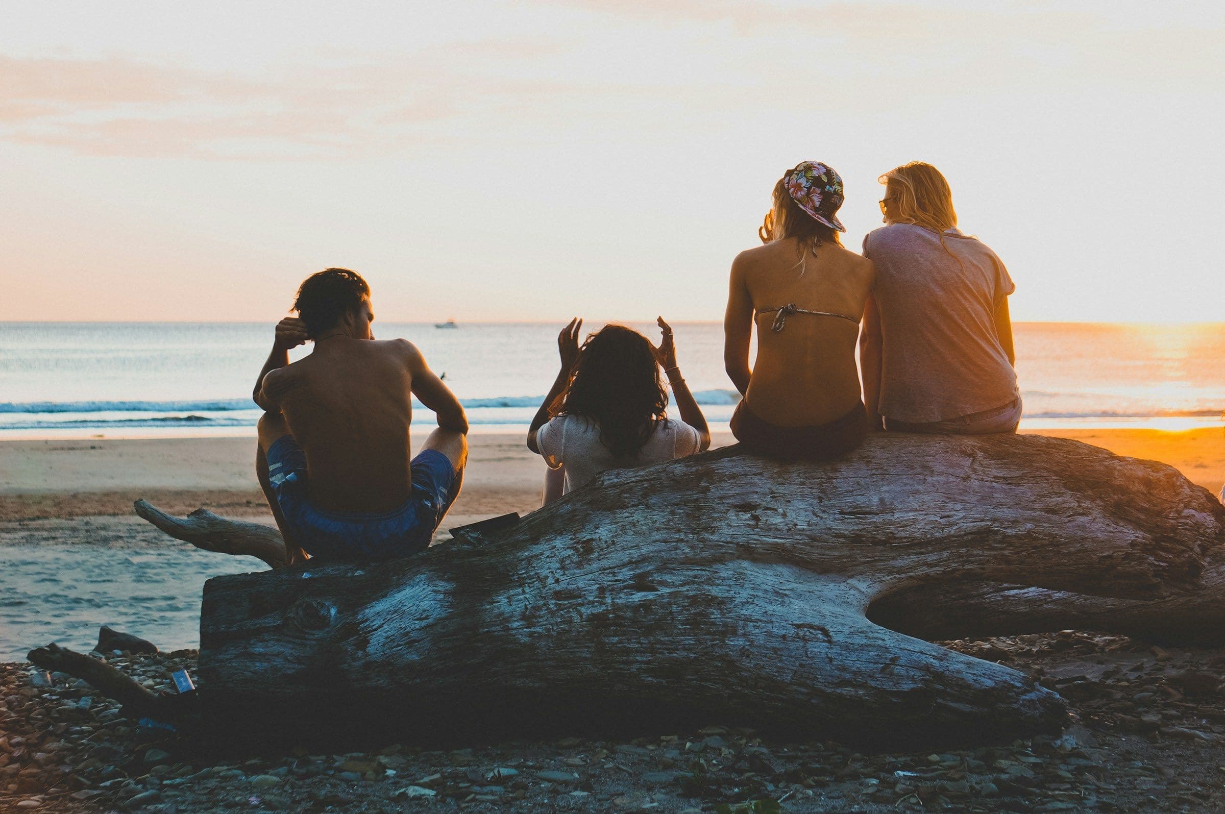 Four young friends, seen from behind, sit on a big driftwood log on a quiet beach, watching the sun set over the sea.