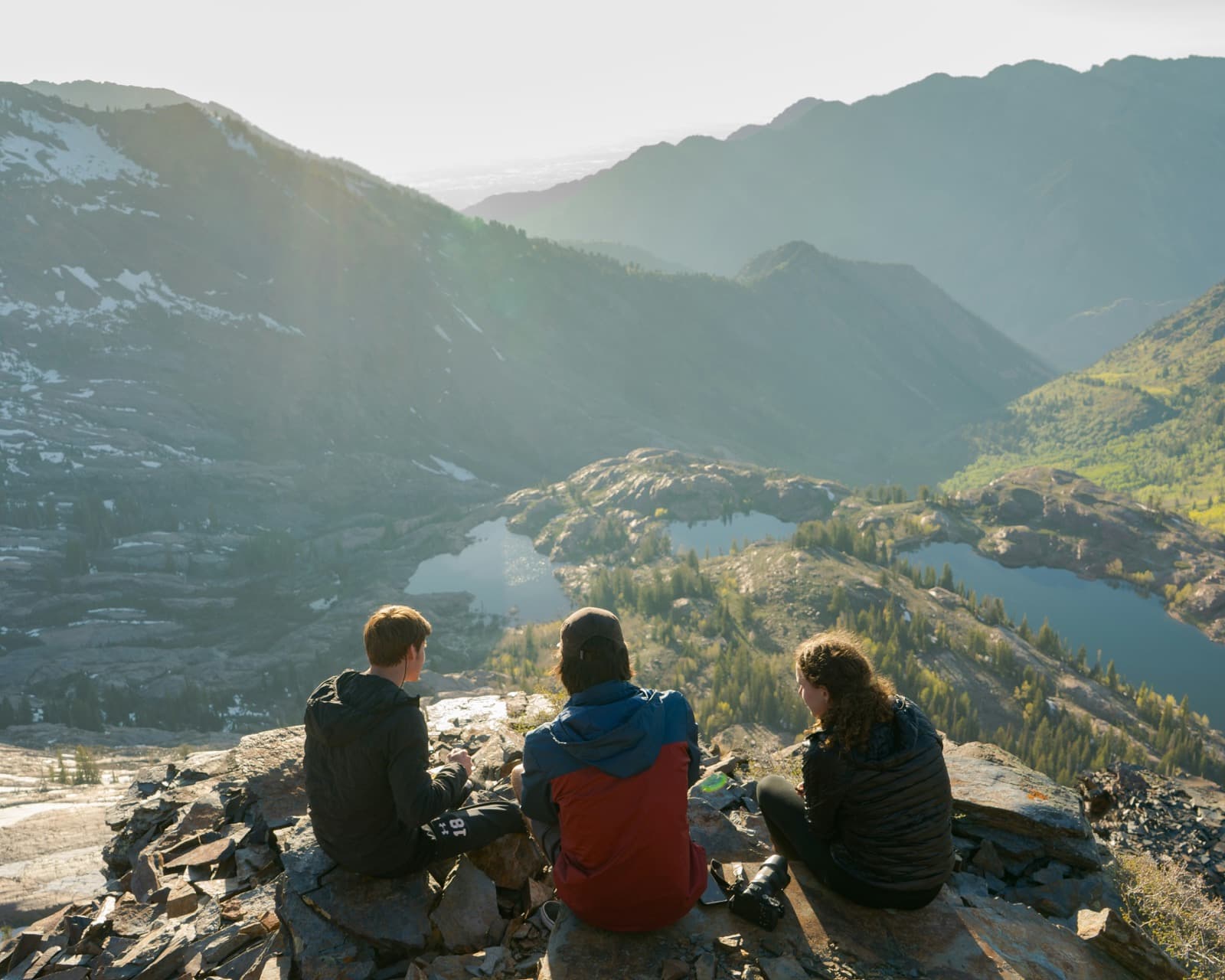 Three young friends sitting on rocks high on a mountain, looking out over alpine lakes and sunlit peaks.