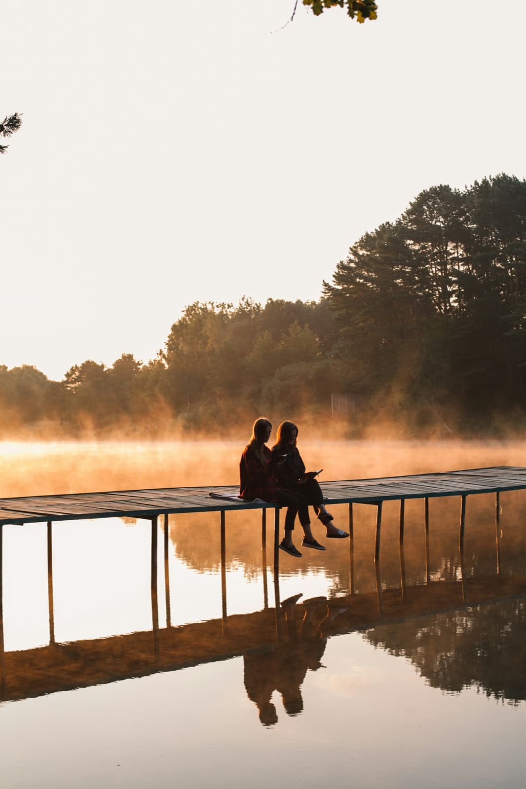 Two young people, seen from a distance, sitting on a long wooden dock over a still lake as morning mist glows gold.