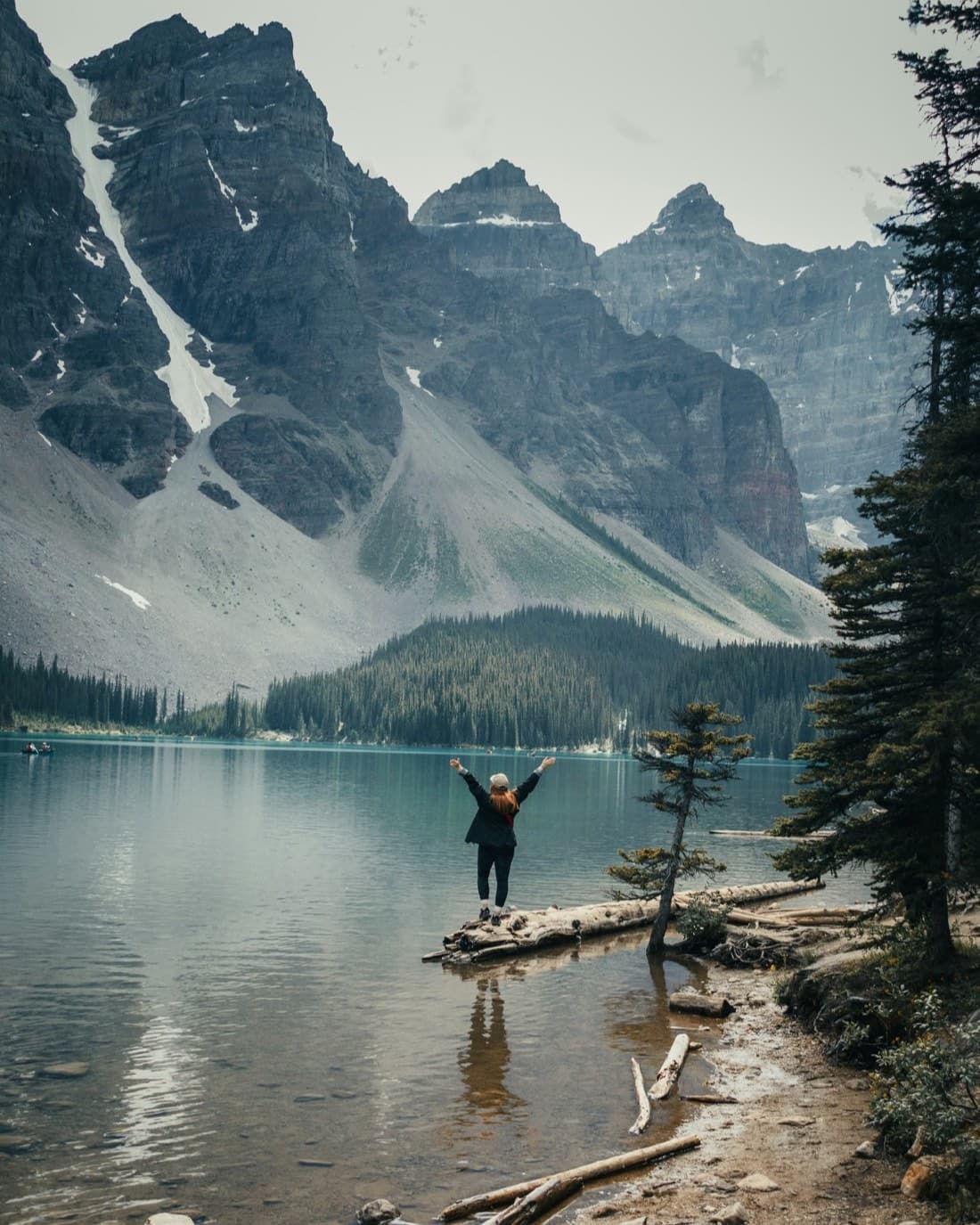 A young hiker with arms raised stands on a fallen log at the edge of a turquoise mountain lake under tall peaks.