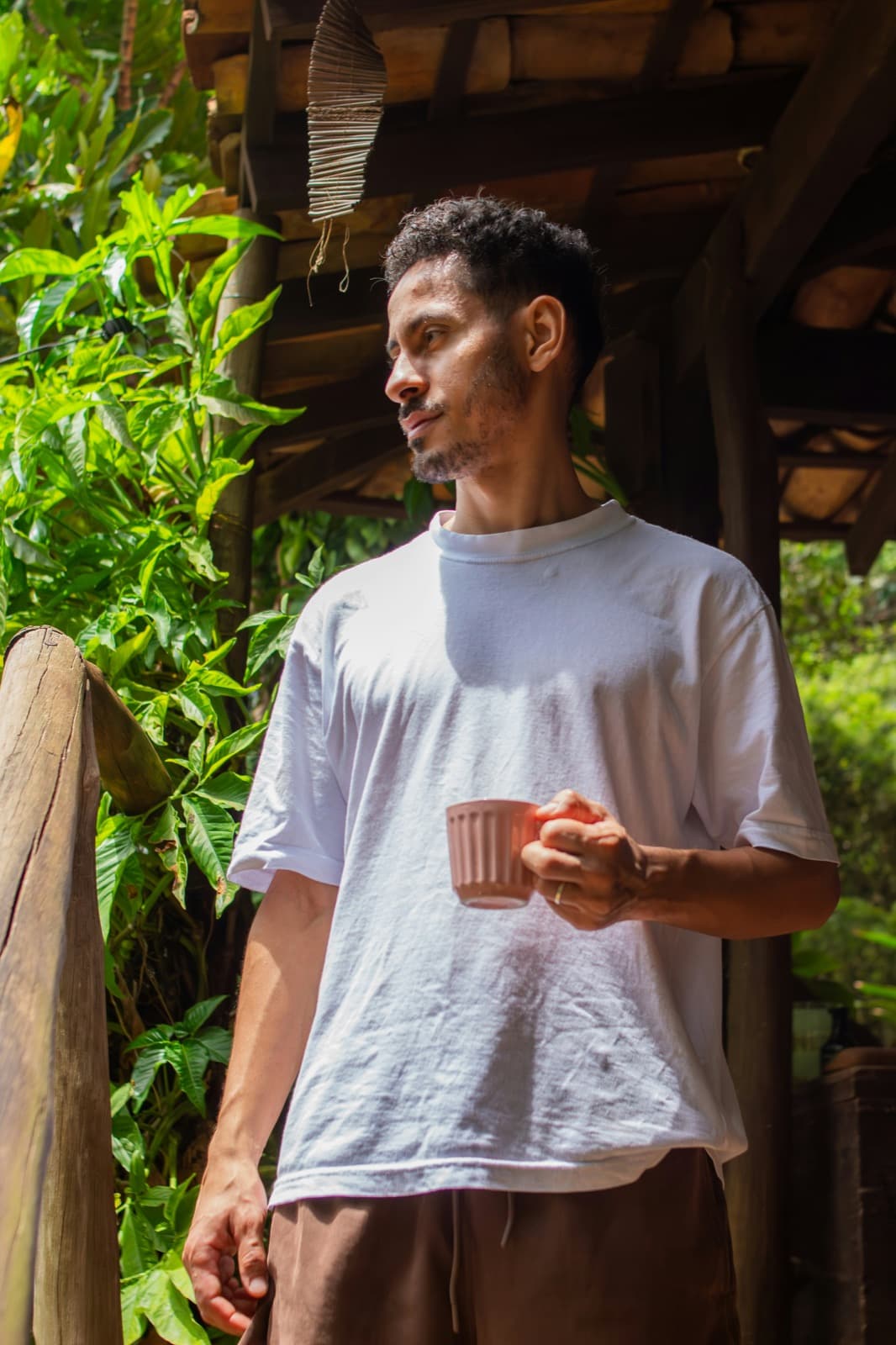 A young man in a white T-shirt holding a mug of coffee on a wooden porch surrounded by green leaves, gazing out.