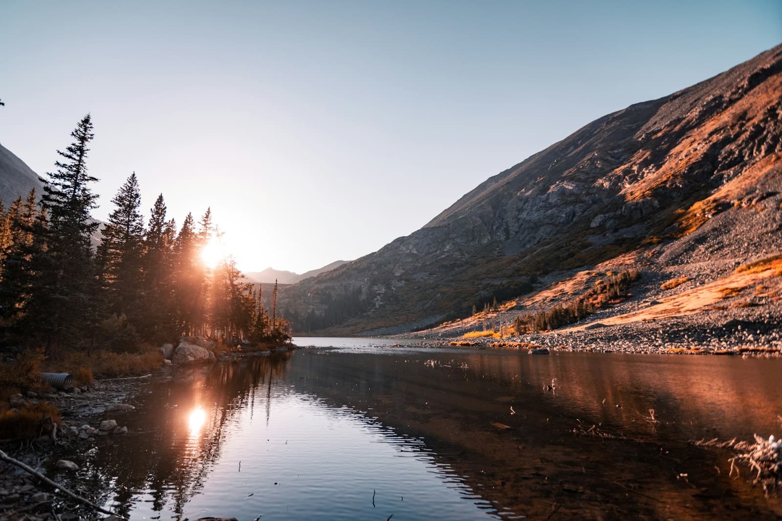 The sun rising over pine trees beside a still mountain lake.