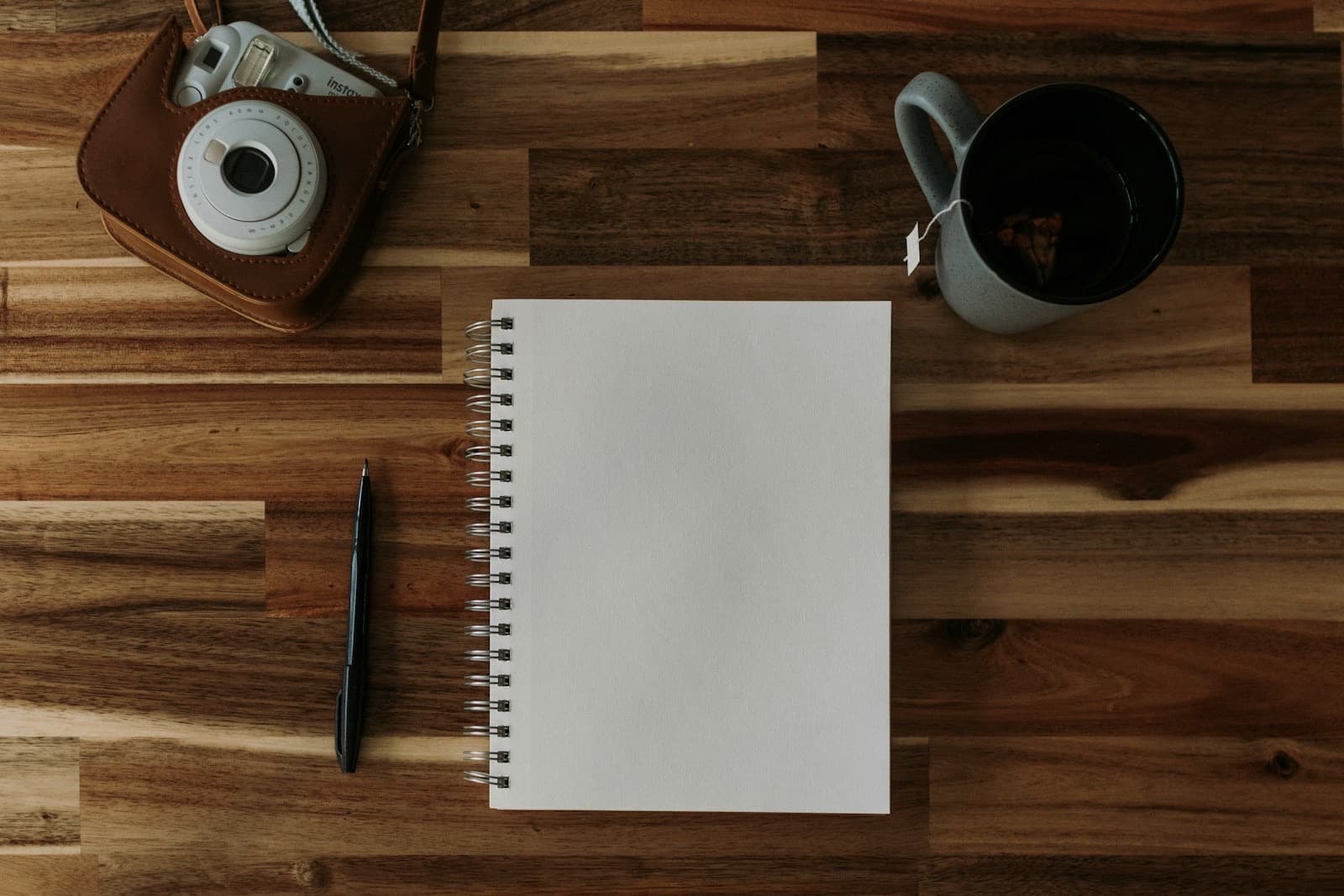 A blank notebook, a pen, a mug and a camera on a wooden table.