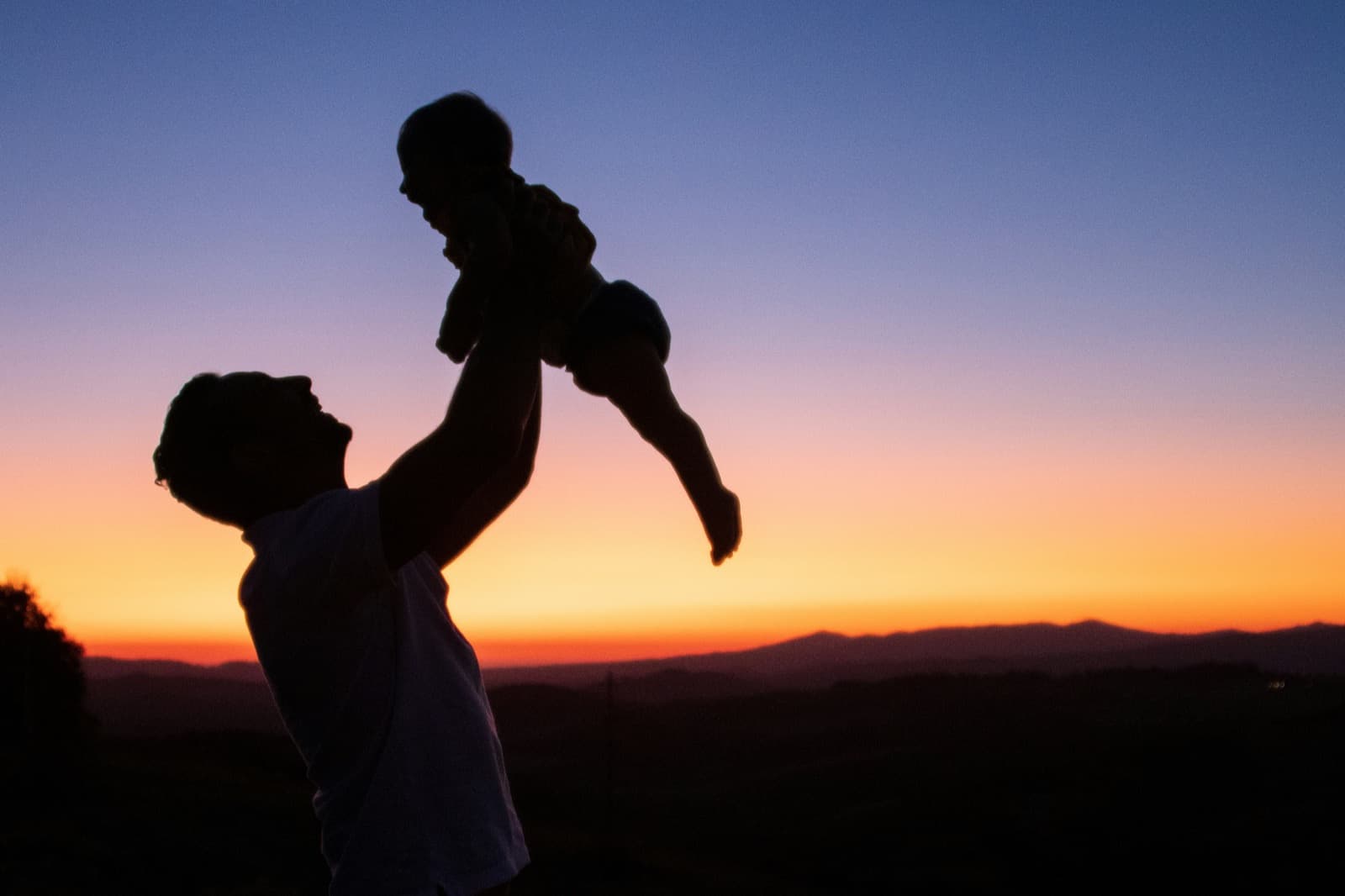 The silhouette of a young parent lifting a baby high into an orange and violet sunset sky above the hills.