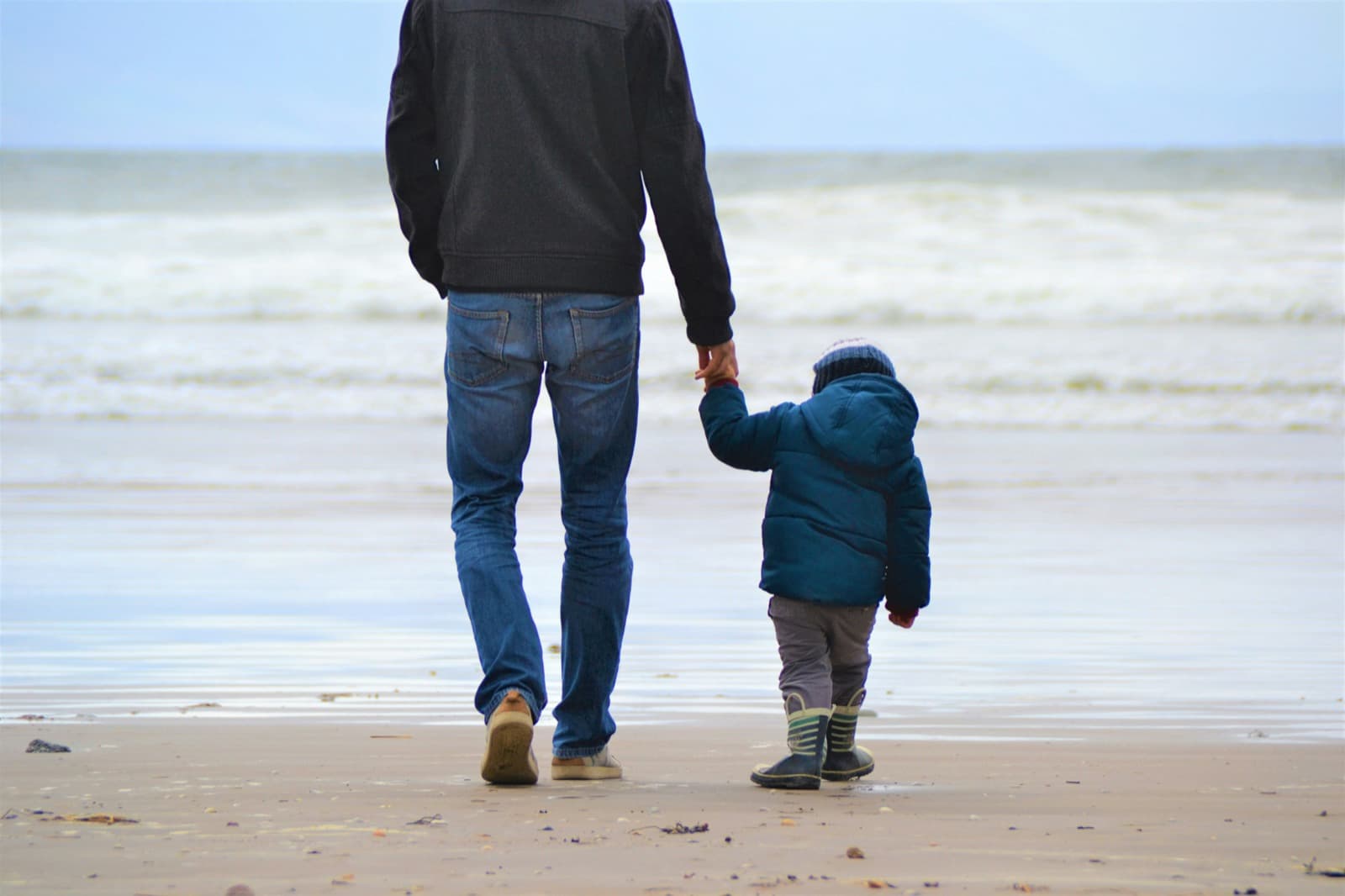 A man and a toddler in rain boots, seen from behind, holding hands as they walk toward the waves.