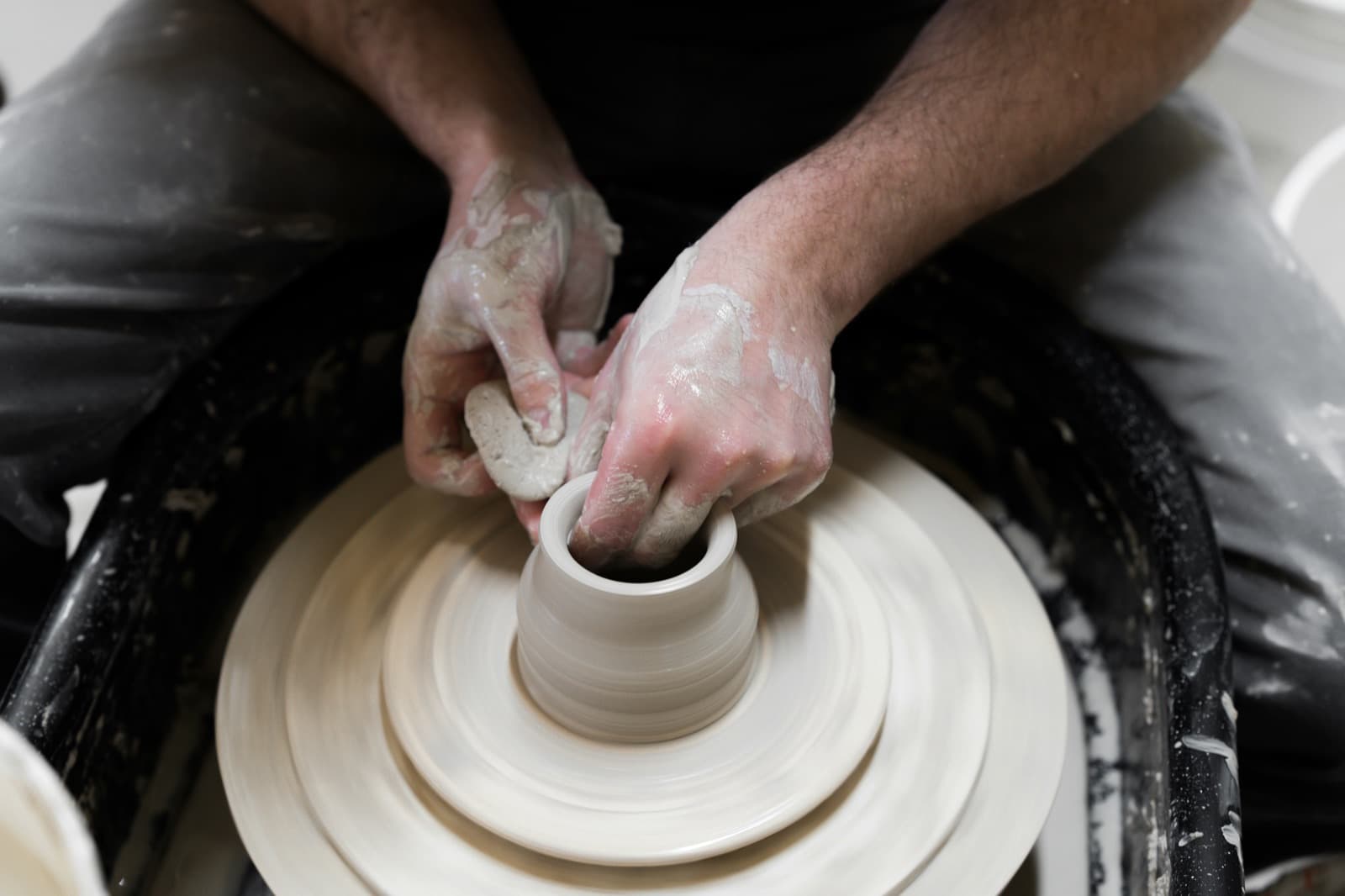Clay-covered hands shaping a small pot on a spinning pottery wheel.