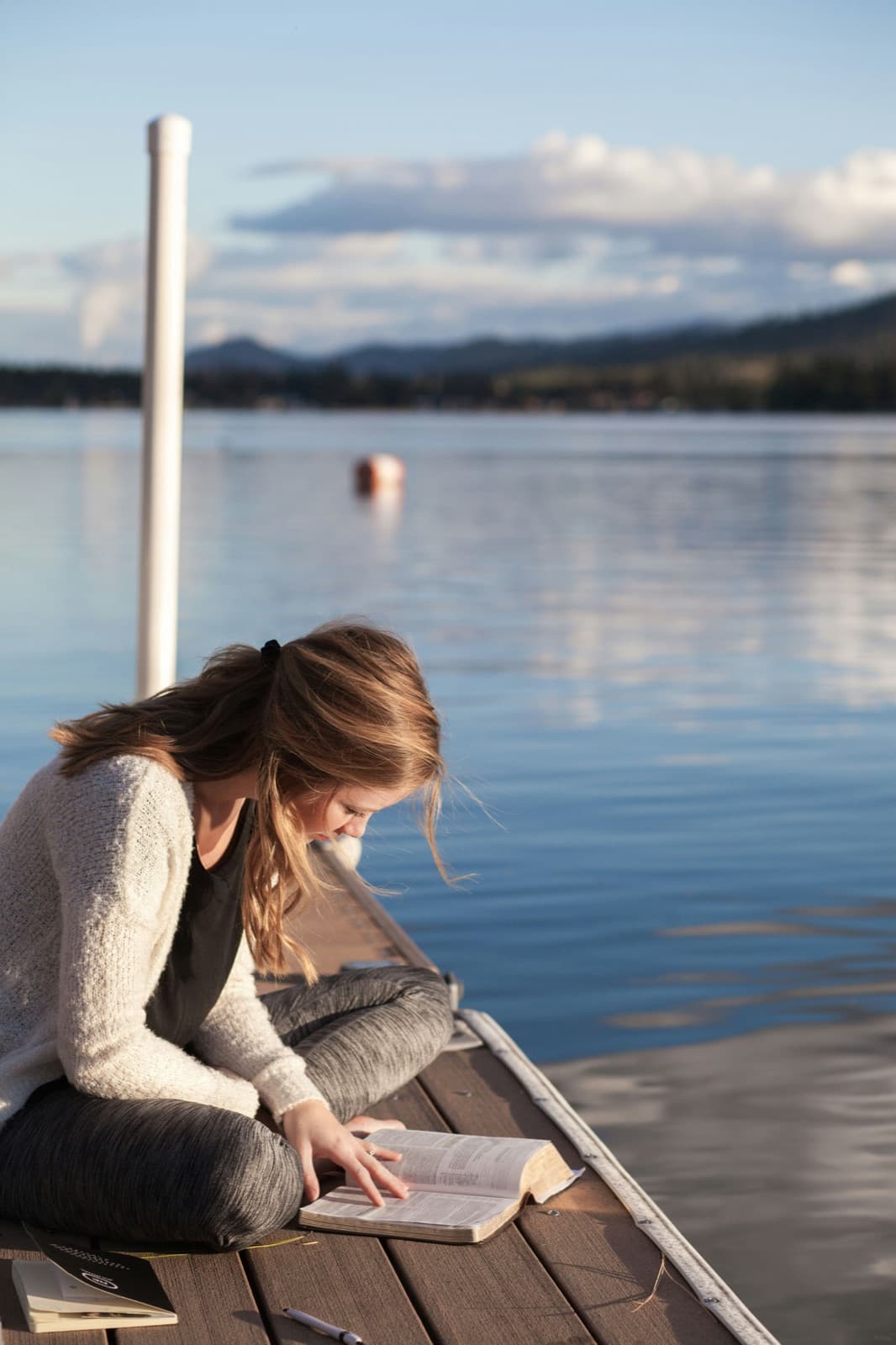 A young woman in a cream cardigan sits cross-legged on a wooden dock, reading a book, with a calm blue lake and hills behind her.
