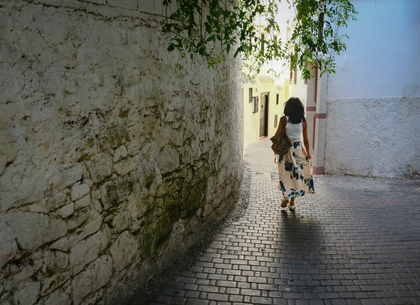 A young woman in a flowered skirt, seen from behind, walking up a sunlit cobblestone lane between old whitewashed walls.