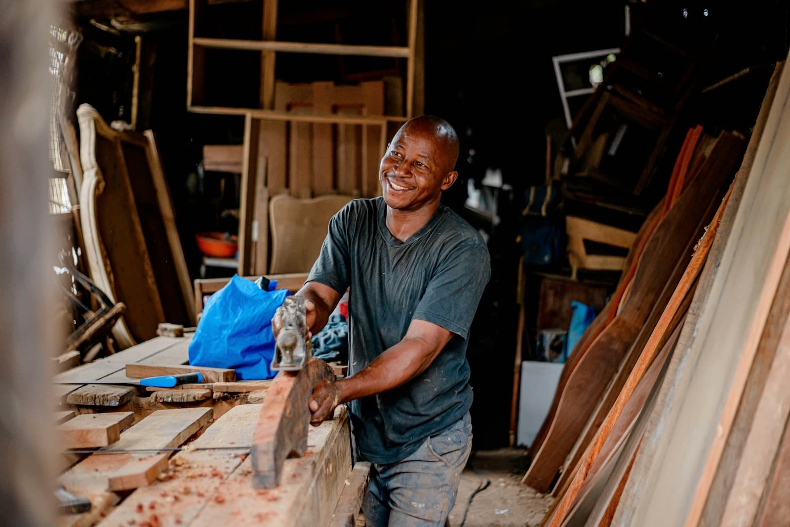 A smiling man planing a board in a woodworking shop full of lumber.